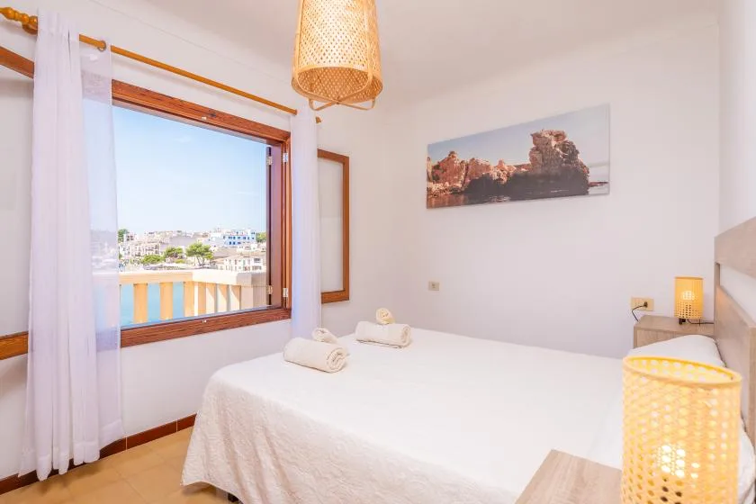 Un dormitorio luminoso con una cama blanca junto a una ventana grande con vistas a edificios.