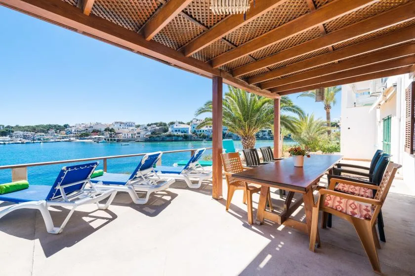 Una terraza frente al agua tiene asientos reclinables y una mesa de comedor bajo una pérgola.