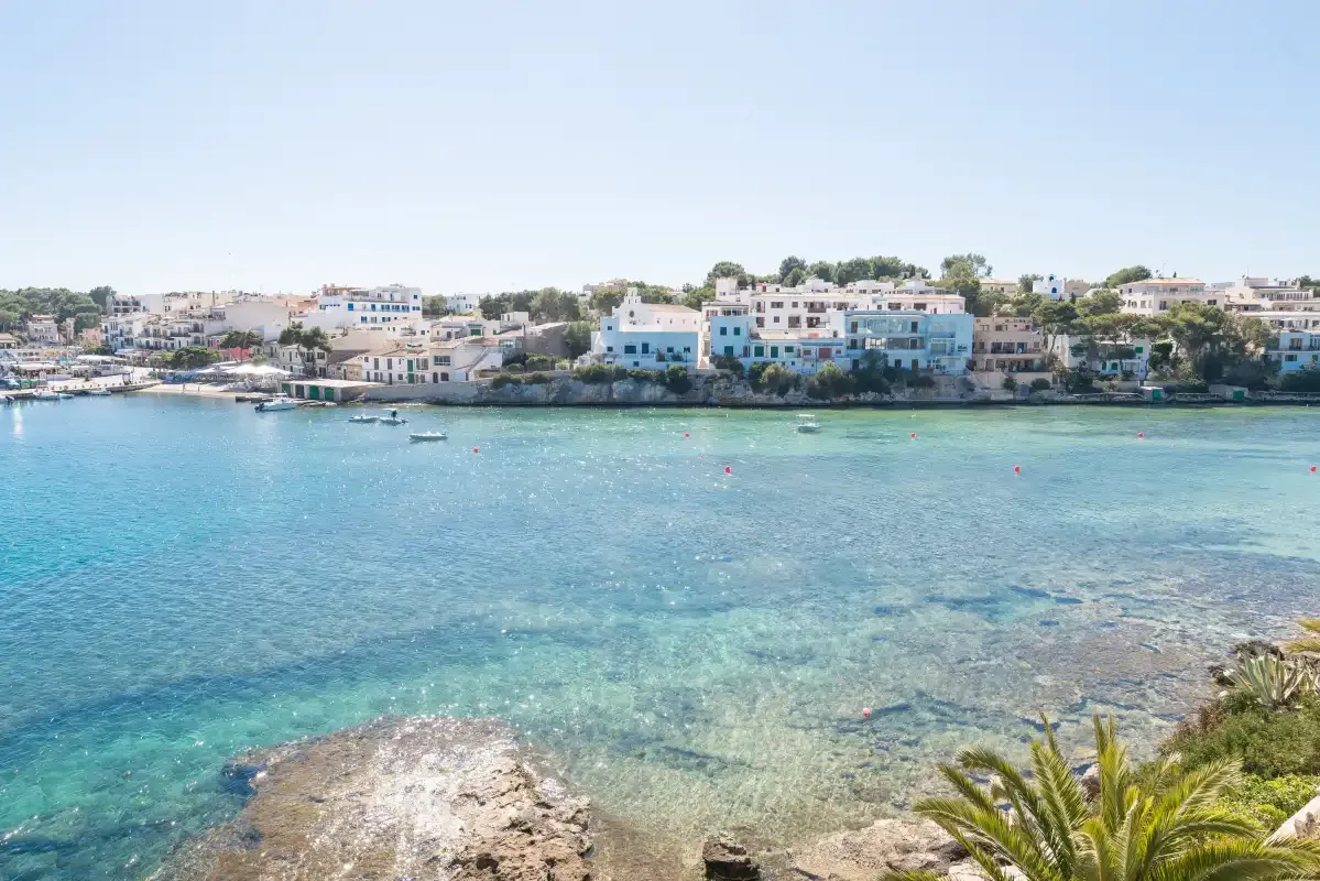 Una bahía costera con aguas turquesas y claras, zonas rocosas poco profundas y edificios junto a la costa.