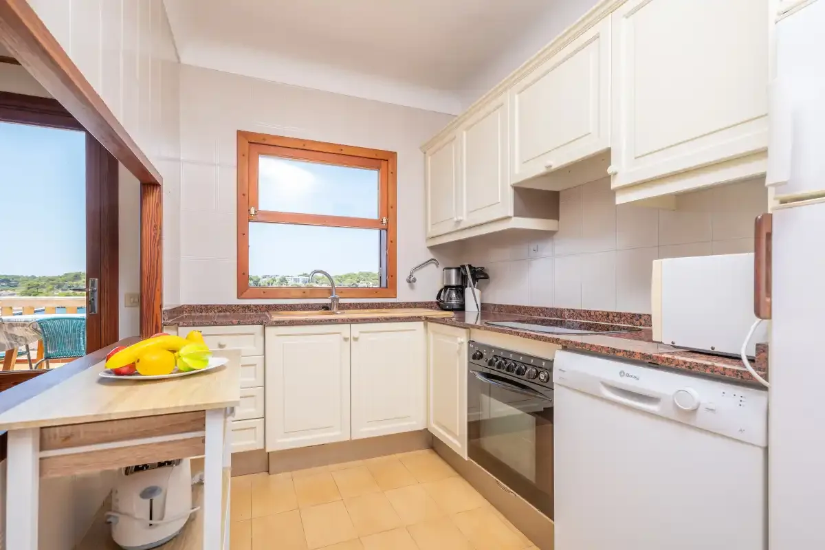 Una cocina luminosa con armarios blancos, suelo de baldosas y una ventana con vistas al exterior.