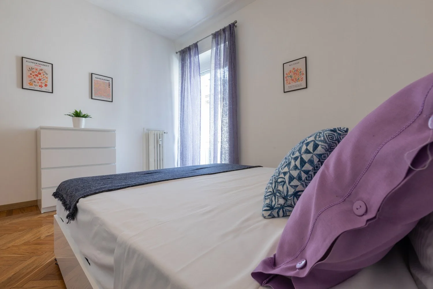 Un dormitorio con una cama bien tendida y una ventana alta con cortinas translúcidas.
