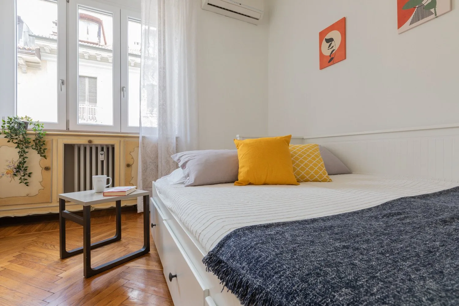 Un dormitorio con una cama, cojines amarillos y ventanas altas.