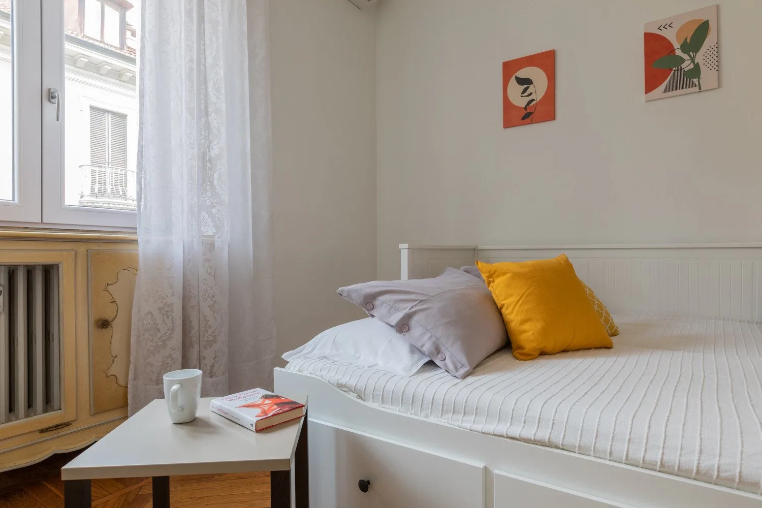 Dormitorio con una cama blanca, cojines de colores y una pequeña mesa junto a la ventana.