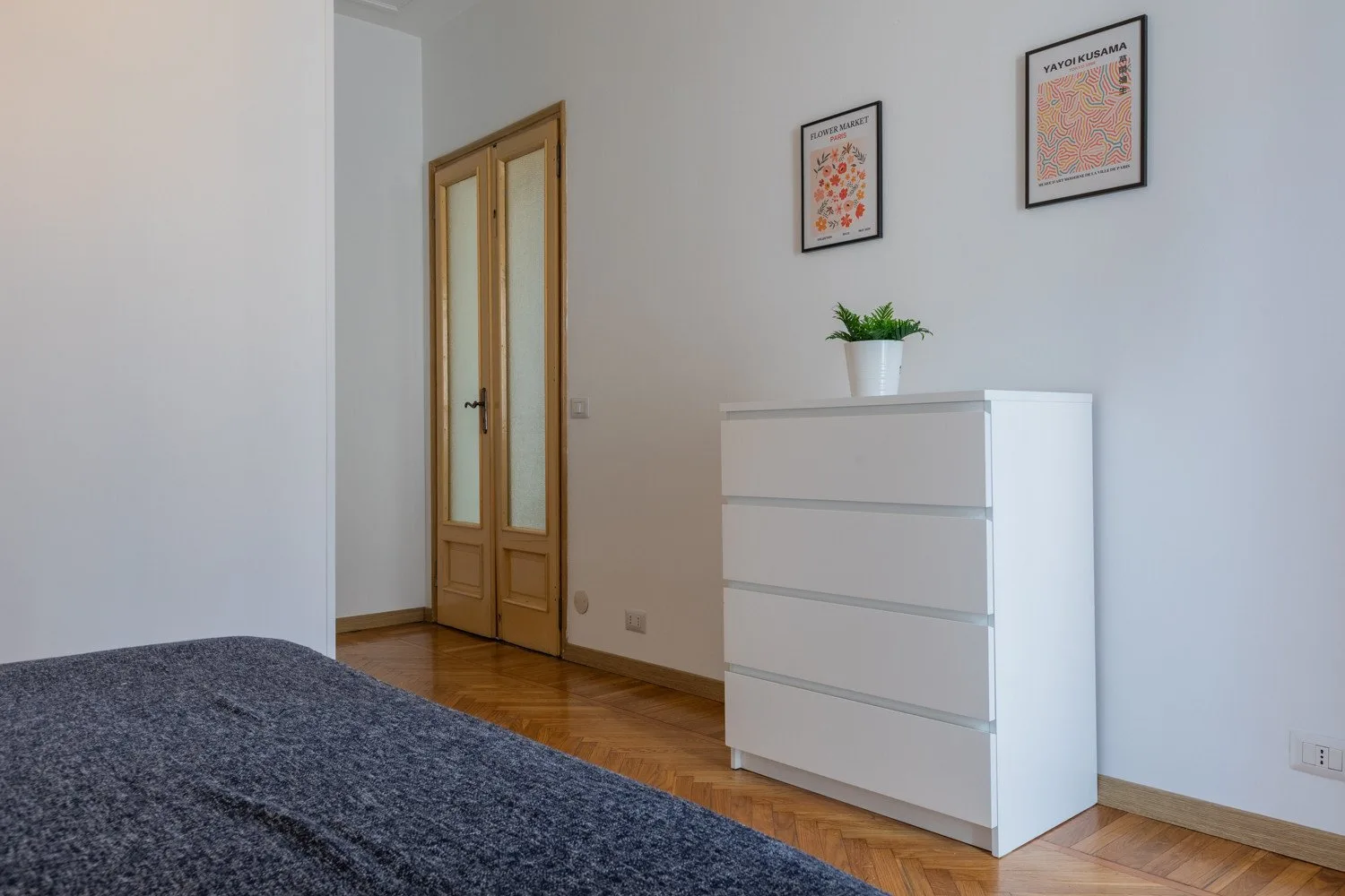 Un dormitorio luminoso con una cómoda blanca, láminas enmarcadas y una puerta de madera.