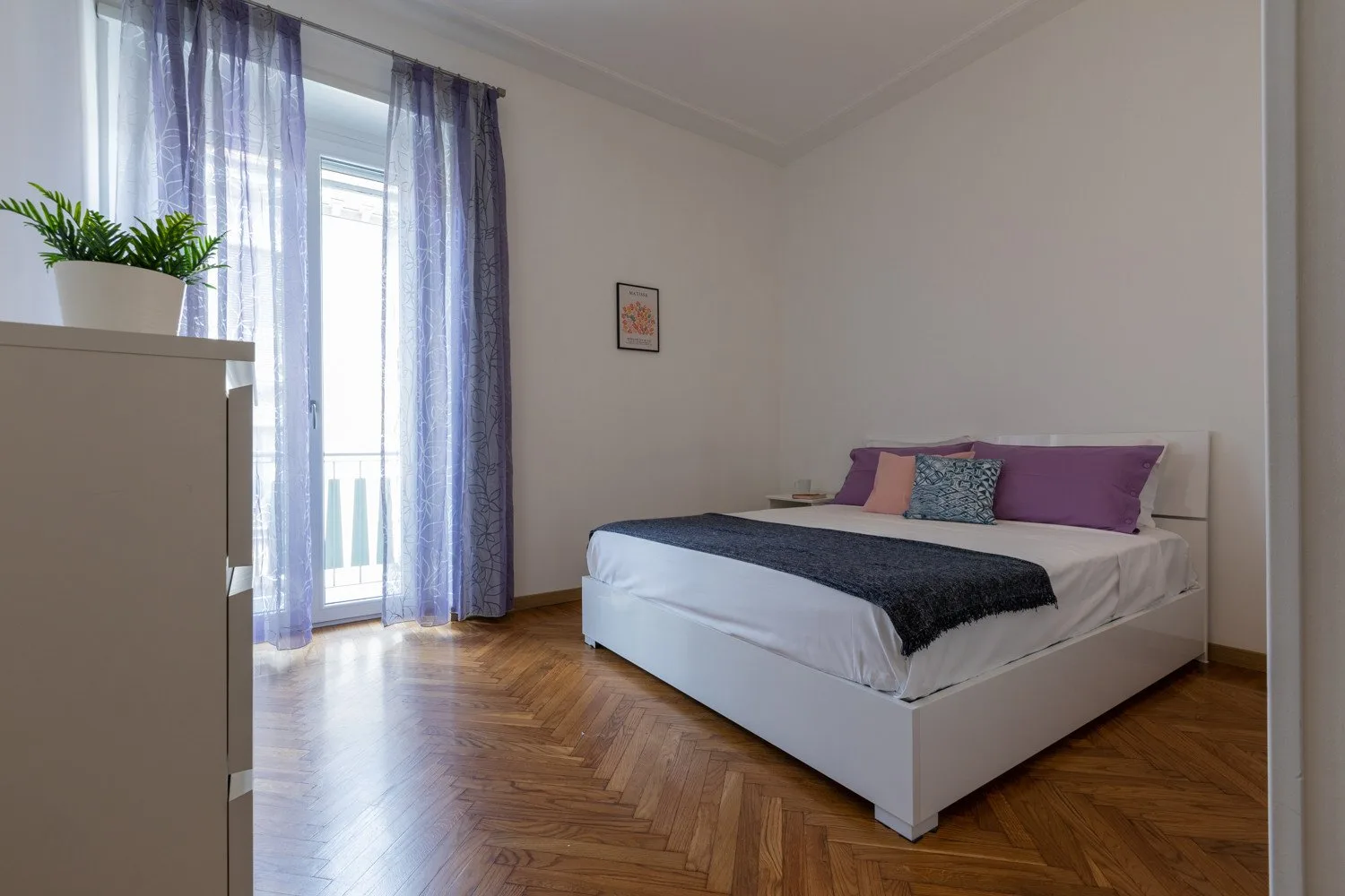 Un dormitorio con una cama blanca, almohadas de colores y cortinas translúcidas junto a la ventana.