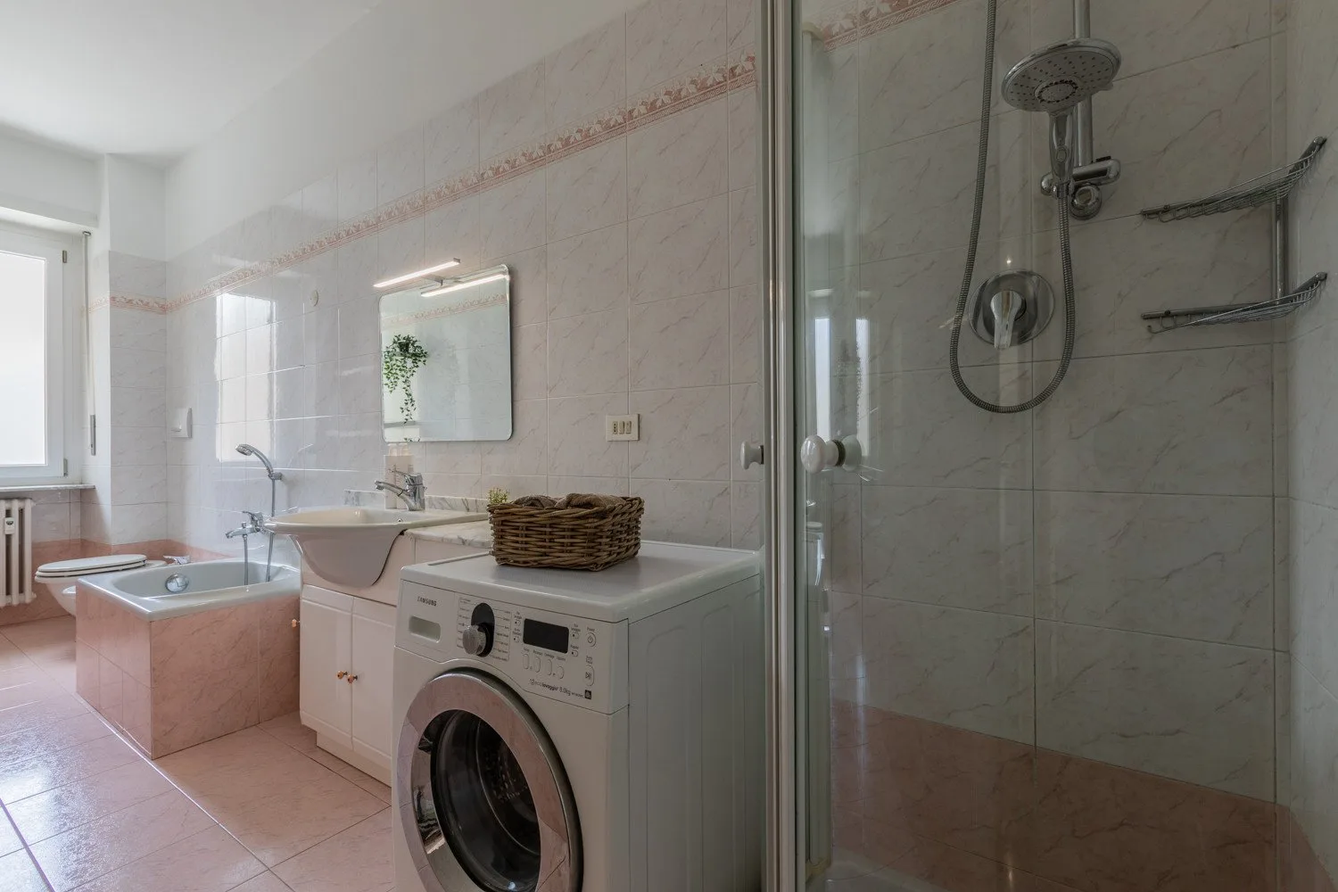 Un baño alicatado con bañera, lavadora y ducha con mampara de vidrio.