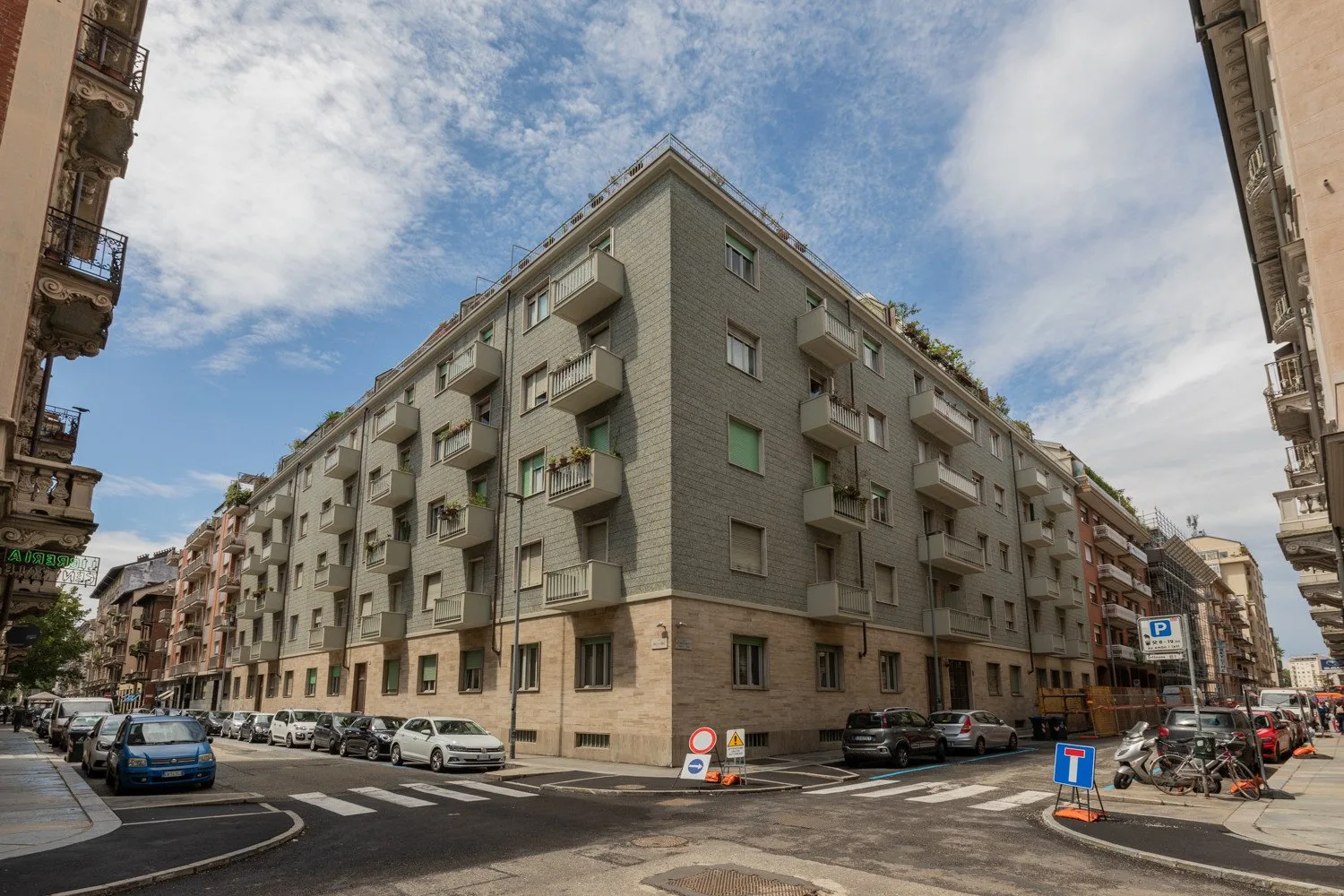 Un edificio de apartamentos de varias plantas ocupa una esquina urbana bajo un cielo parcialmente nublado.