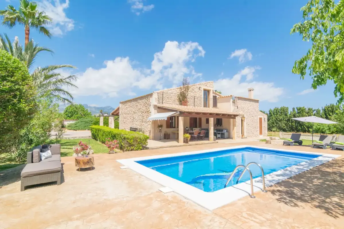 Una piscina está junto a una casa de piedra, con asientos de descanso y vegetación cerca.