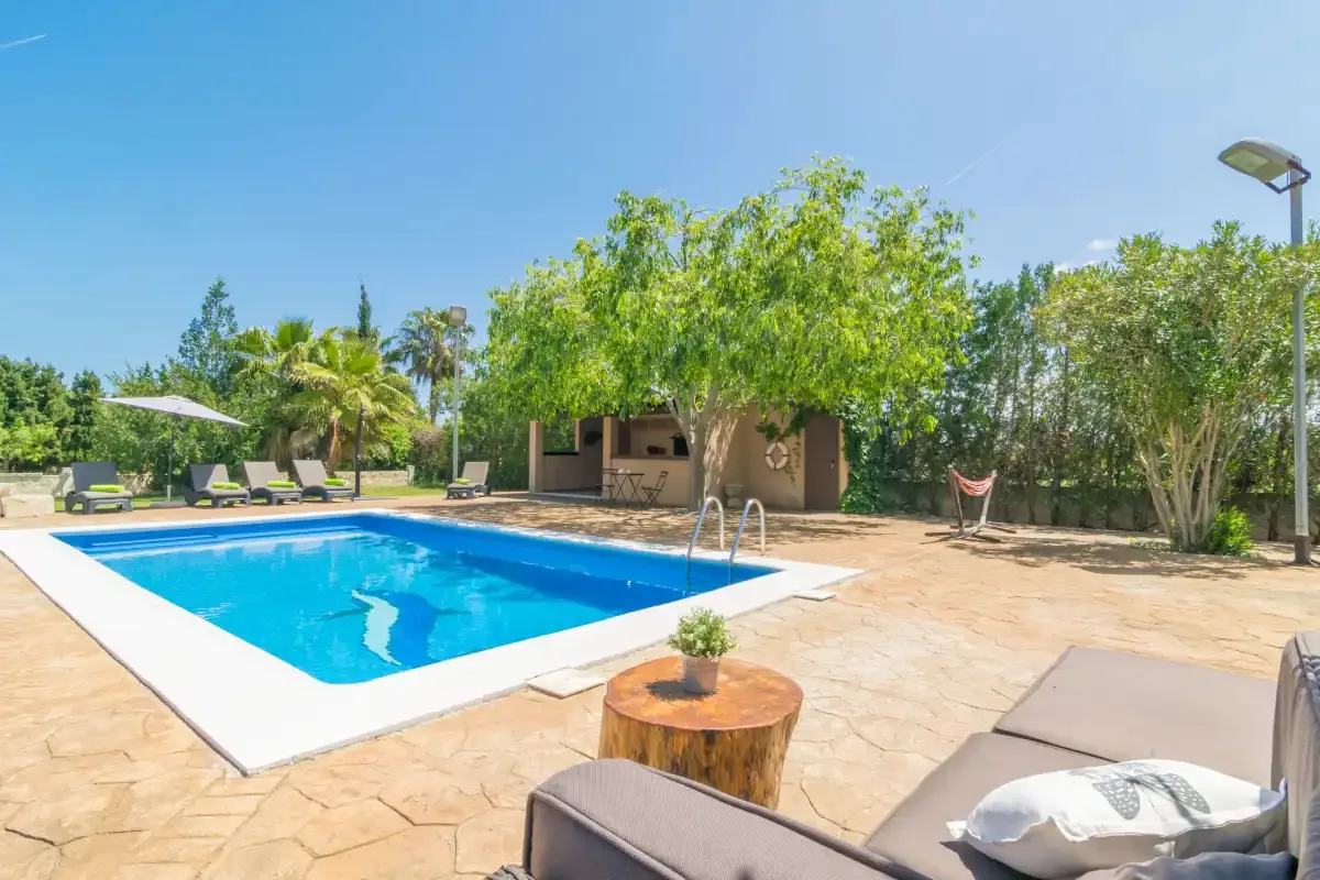 Una piscina al aire libre está junto a un patio pavimentado con asientos y árboles frondosos.
