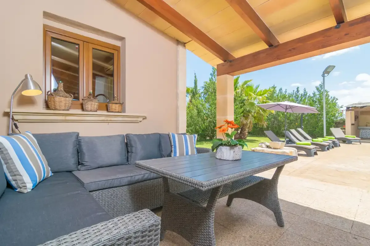 Un patio cubierto tiene asientos y una mesa al lado de un jardín soleado.