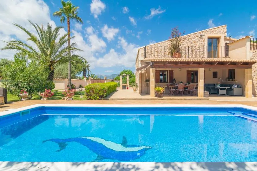 Una piscina frente a una casa de piedra con una zona de estar a la sombra.