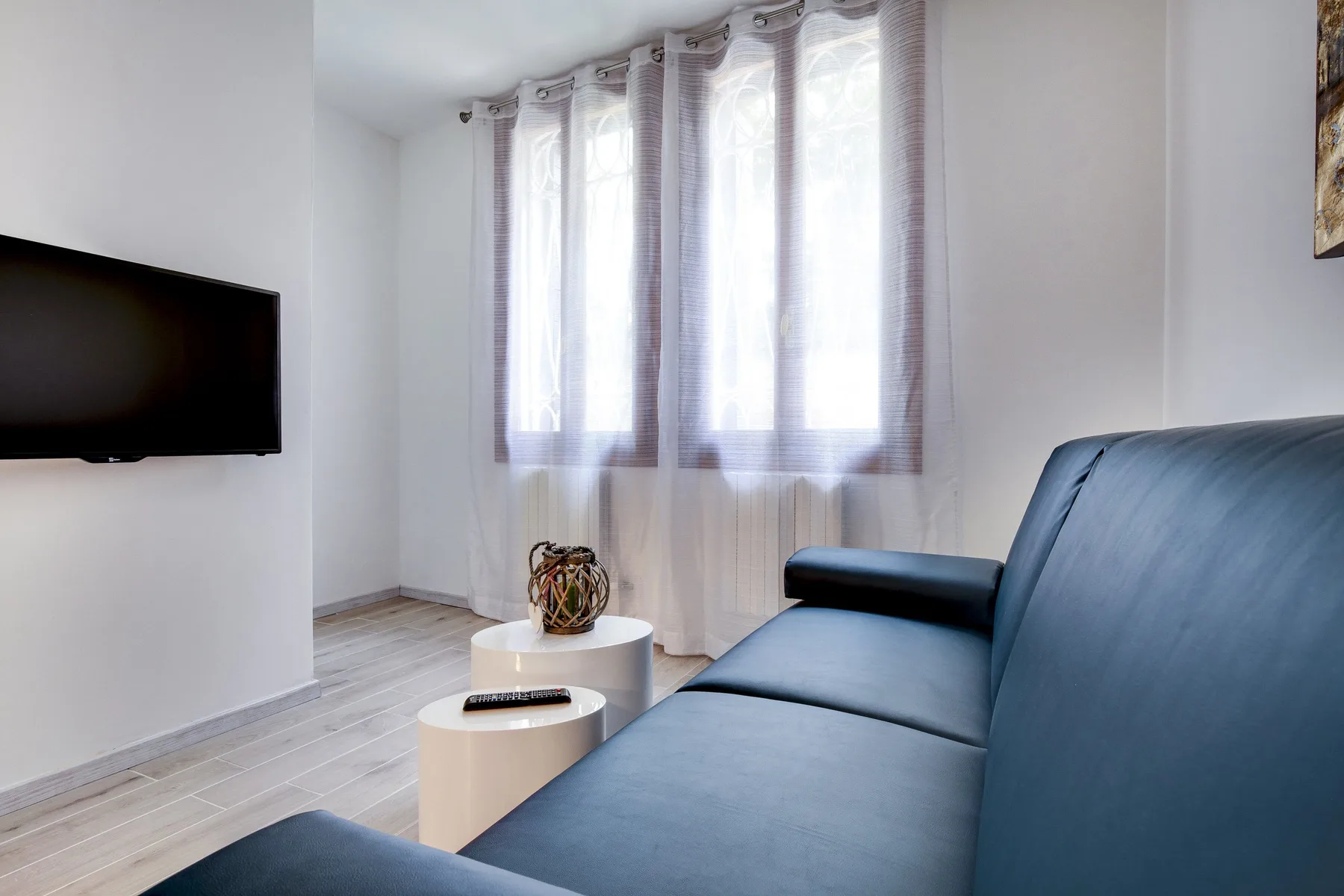 Una sala luminosa con un sofá azul, un televisor y ventanas altas con cortinas.