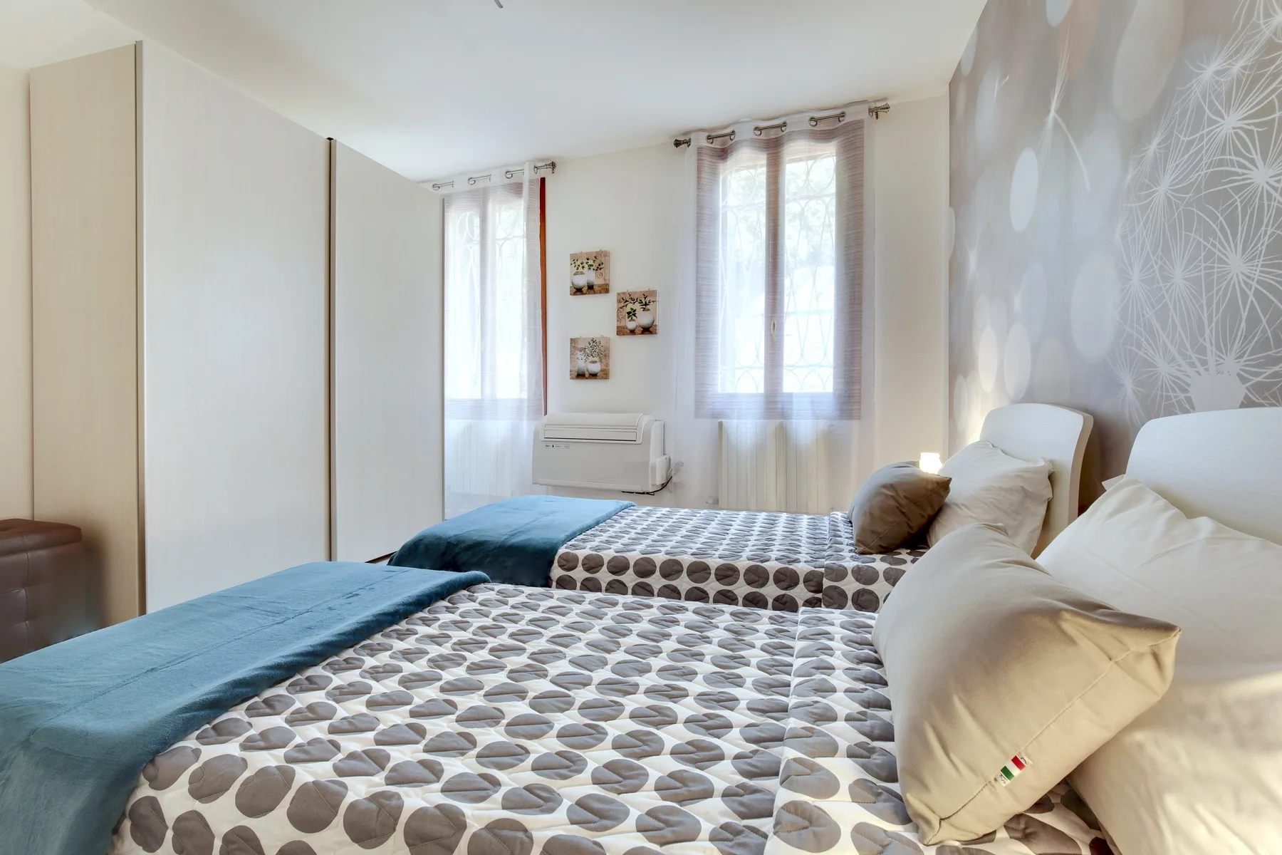 Un dormitorio con dos camas, ropa de cama estampada y ventanas con cortinas claras.