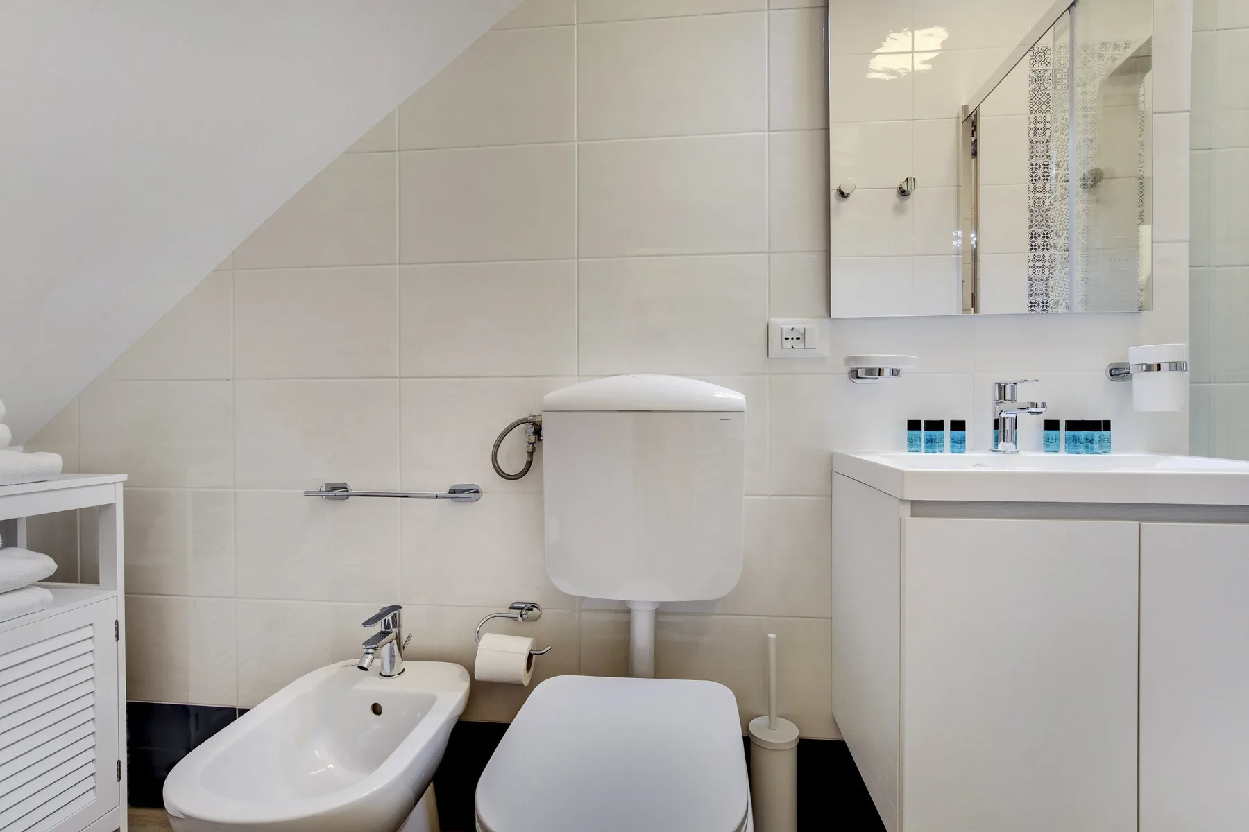 Un baño con azulejos, inodoro, bidé y lavabo bajo un techo inclinado.