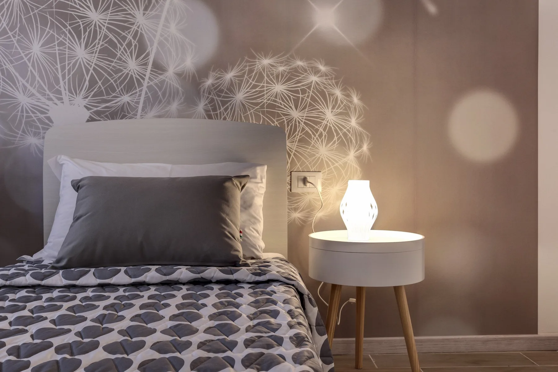 Un dormitorio con ropa de cama estampada, una mesita y una lámpara encendida.