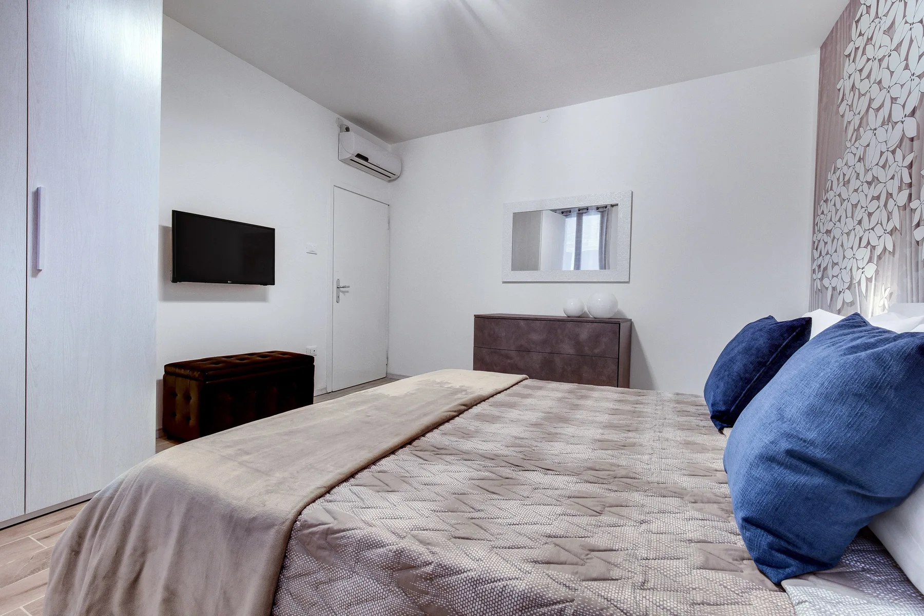 Un dormitorio con una cama tendida, un televisor montado en la pared y una cómoda pequeña.