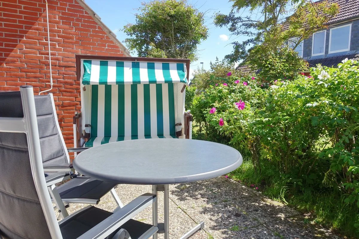 An outdoor patio has a round table, seating, and a striped canopy beside flowering shrubs.