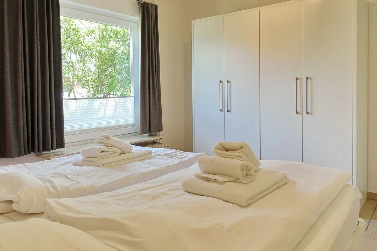 A bedroom with white beds, folded towels, and a large wardrobe beside the window.