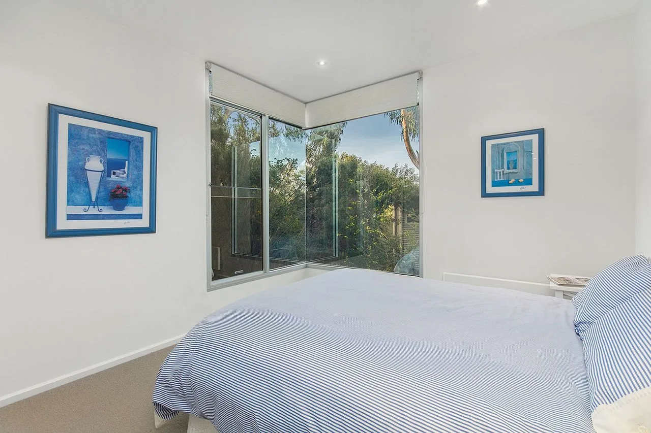 A bright bedroom with a bed beside corner windows overlooking greenery.