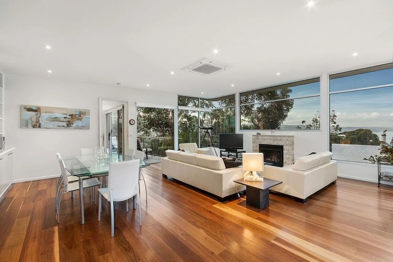 A bright living room with seating, a fireplace, and large windows overlooking greenery.