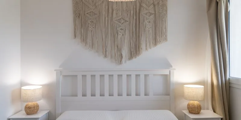 A bedroom with a white bed, bedside tables, and a woven wall hanging.