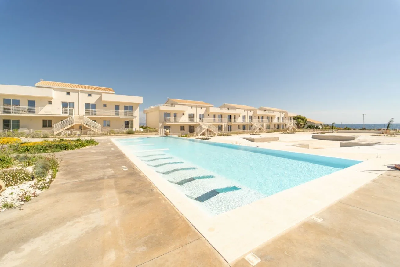 A swimming pool sits in front of a row of low-rise buildings under a clear sky.