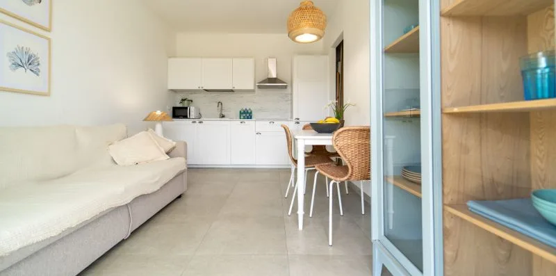 A bright living and dining area opens onto a compact kitchen, with shelving in the foreground.