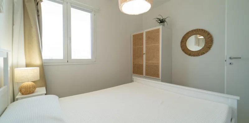 A bright bedroom with a bed, tall windows, and a woven wardrobe.