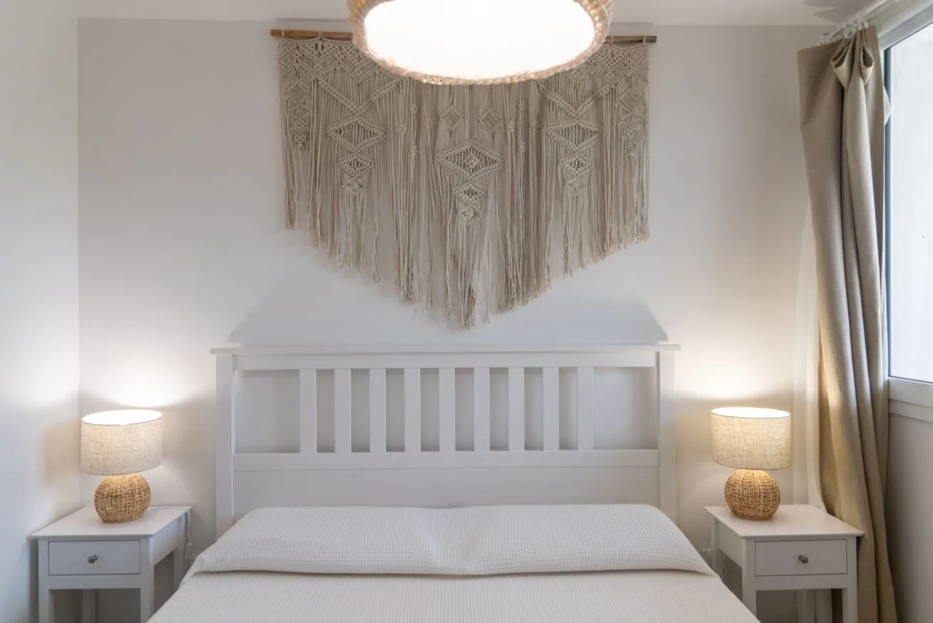 A bedroom with a white bed, matching bedside tables, and a woven wall hanging.