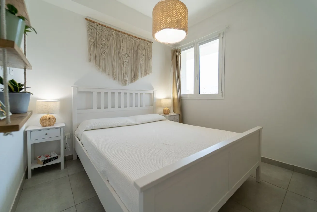 A bright bedroom with a white bed beside a window.