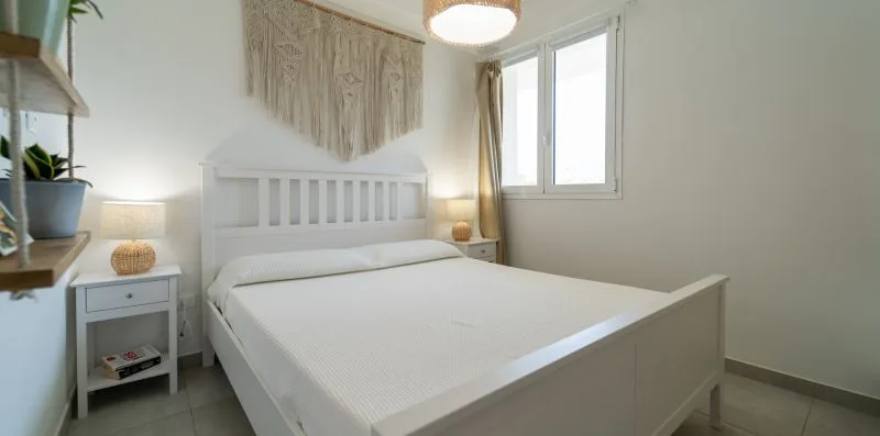 A bright bedroom with a white bed beside a large window.