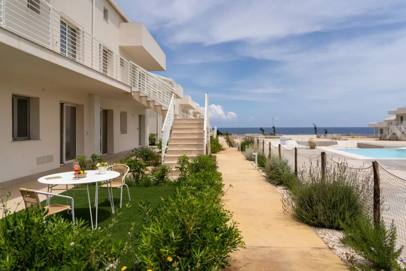 A seaside walkway runs past a white building toward a pool and the ocean.