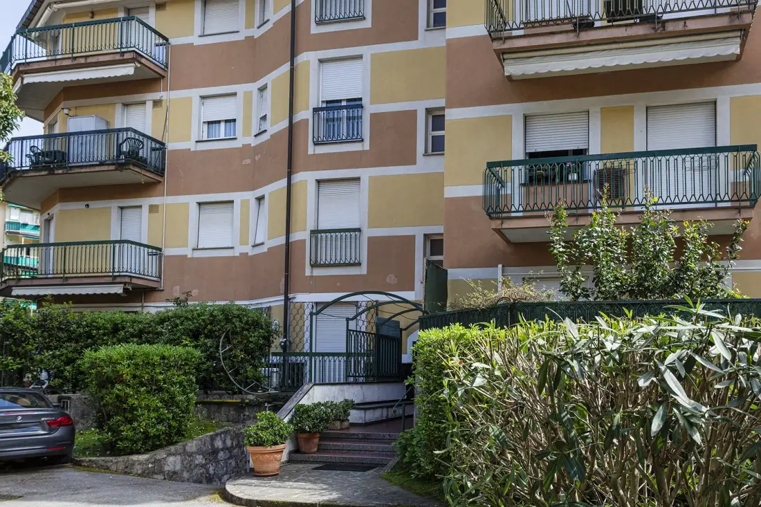 Un edificio residencial de varias plantas con balcones se alza detrás de setos y una entrada con reja.