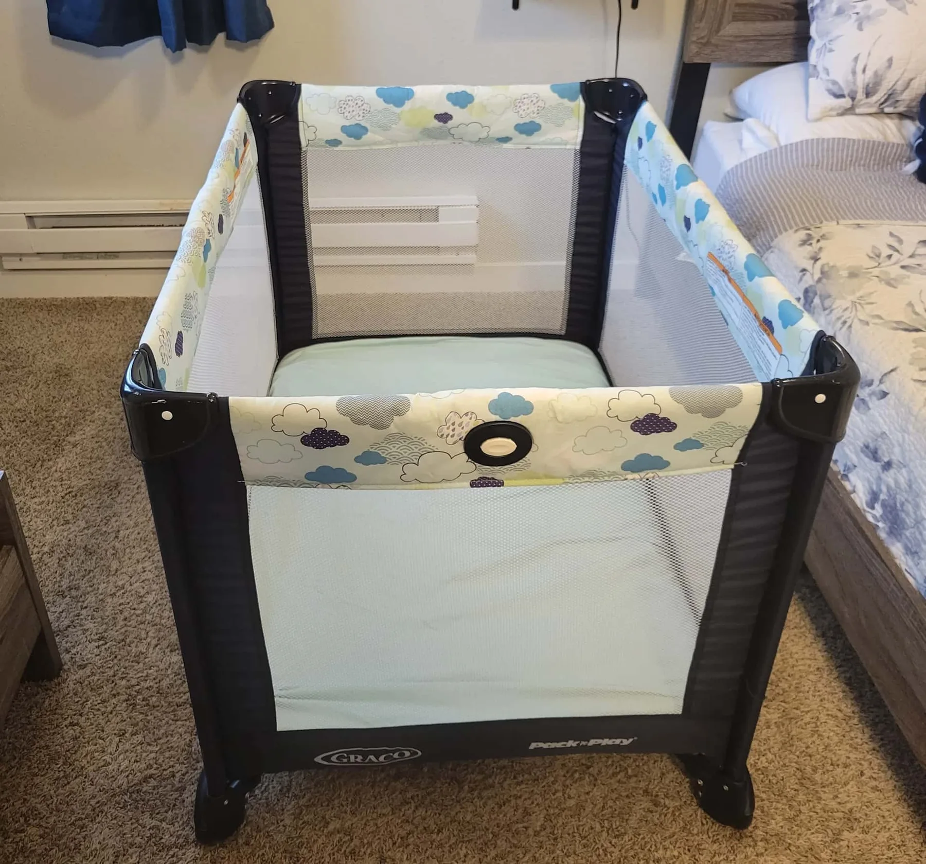 A portable playpen with patterned fabric panels sits beside a bed.