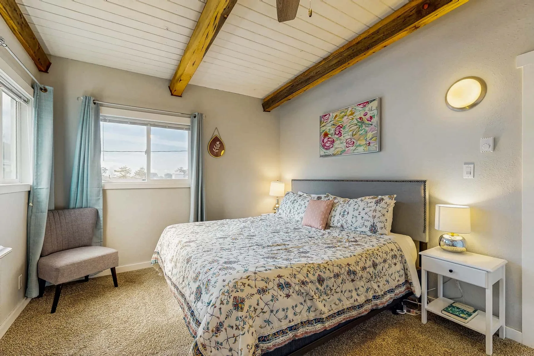 A bedroom with a patterned bedspread, windows, and exposed ceiling beams.