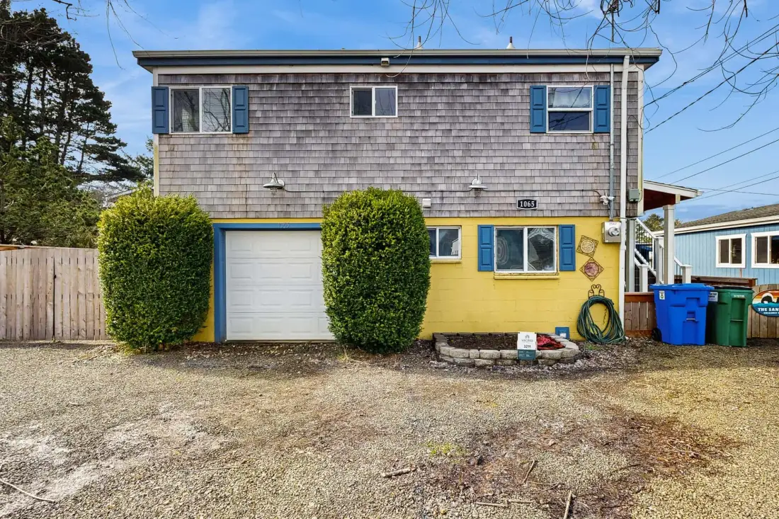 A two-story house with a garage door and shrubs beside the driveway.