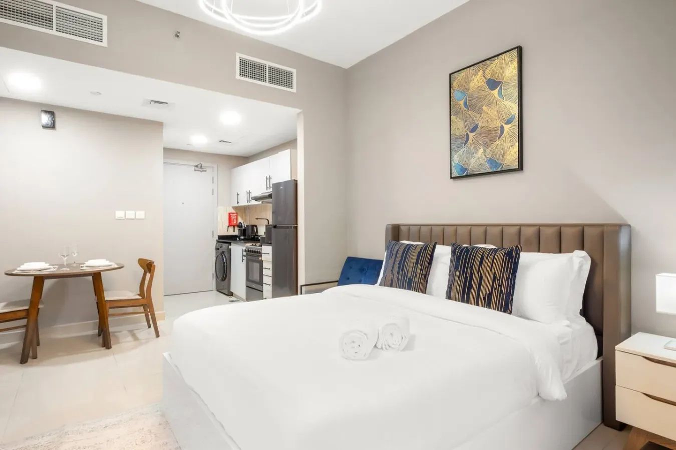 A bright bedroom with a neatly made bed and a small dining area beside the kitchenette.
