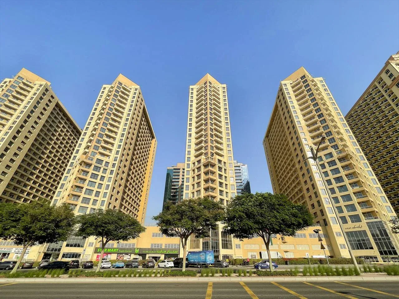 Tall apartment buildings rise above a tree-lined street under a clear blue sky.