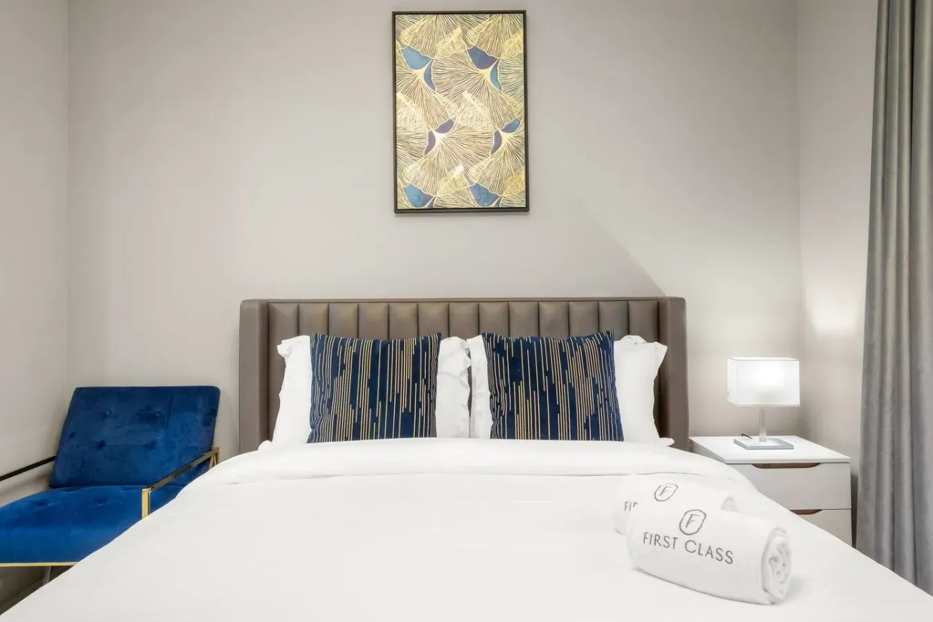 A bedroom with a neatly made bed, blue accent chair, and framed artwork.
