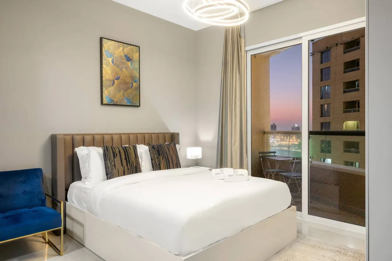 A bedroom with a neatly made bed beside large windows overlooking nearby buildings.