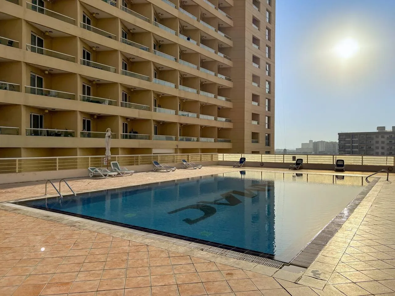 An outdoor swimming pool sits beside a tall building under a bright sky.