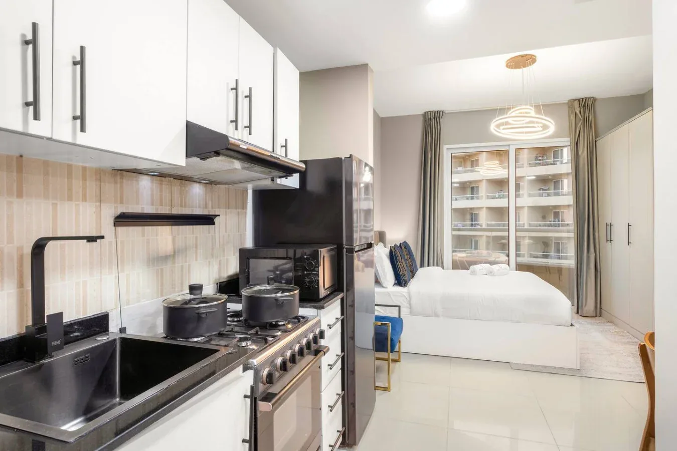A studio room combines a compact kitchen with a bed beside large windows.