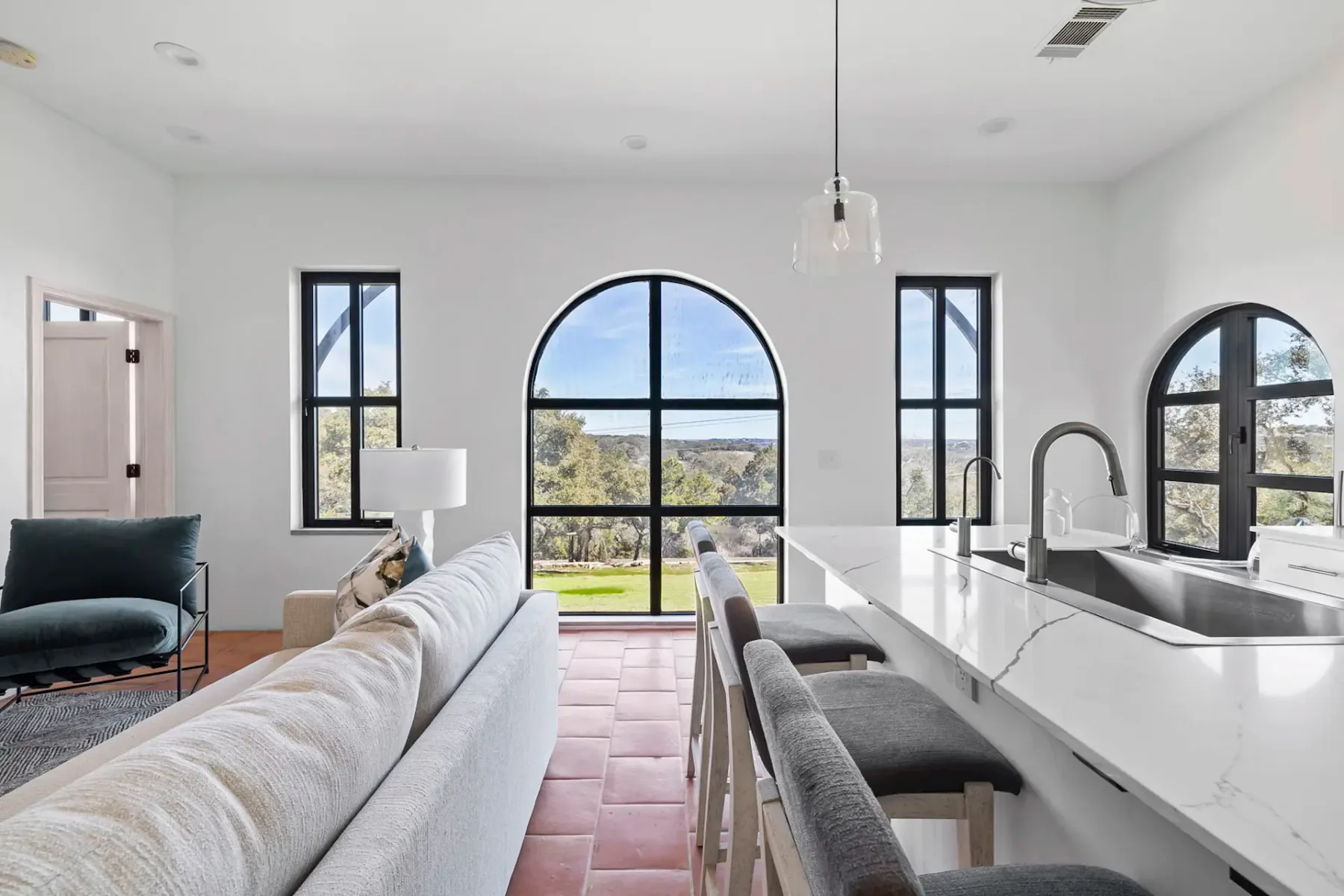Sala de estar moderna con ventanas arqueadas, paredes blancas, sofá, lámpara colgante e isla de cocina con taburetes, con vistas a un paisaje exterior.