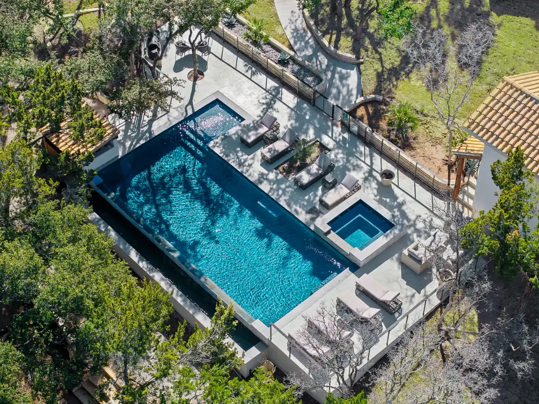 Aerial view of a rectangular swimming pool with adjacent hot tub, surrounded by lounge chairs and greenery.