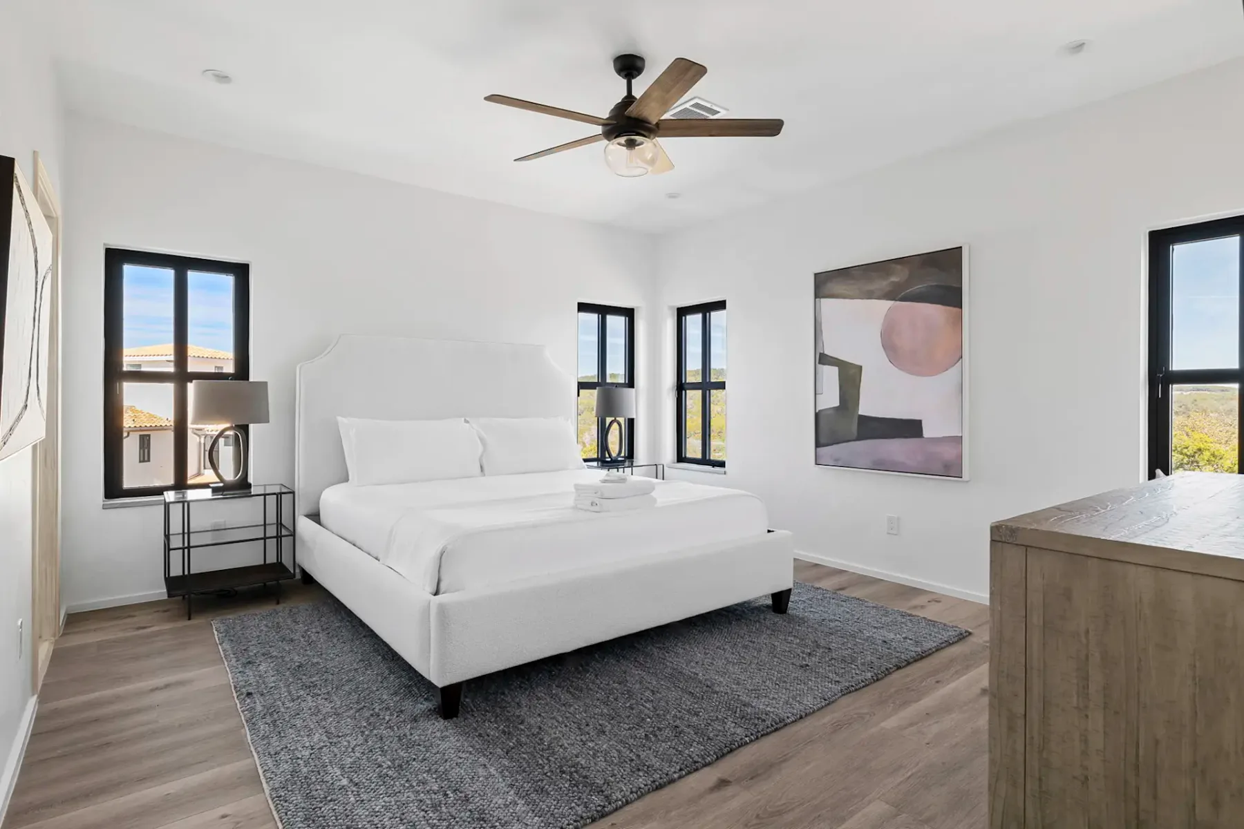 Modern bedroom with a white bed, three windows, a large abstract painting, wooden dresser, and ceiling fan. Neutral tones and natural light.