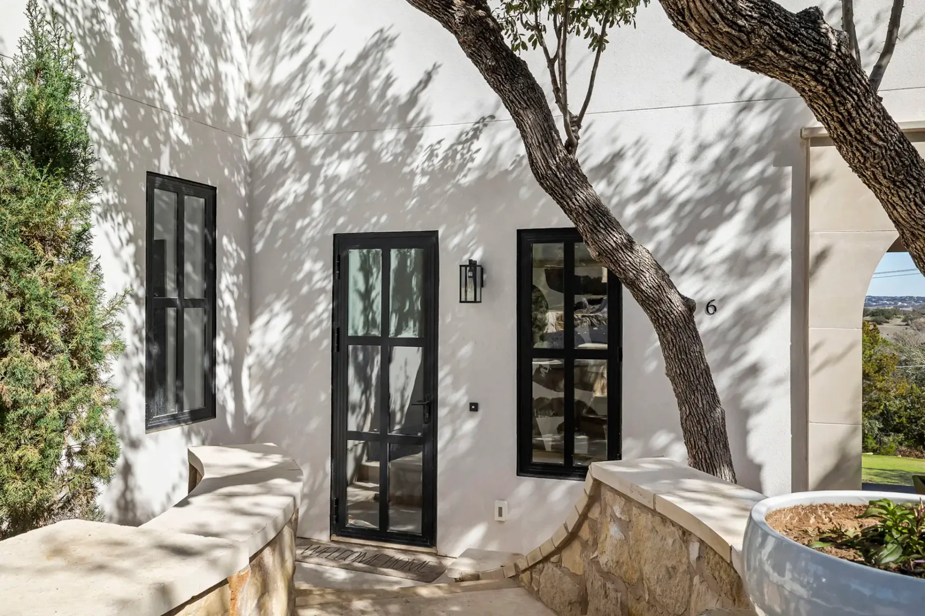 Modern white building exterior with black-framed windows and door, surrounded by stone walls and trees casting shadows.