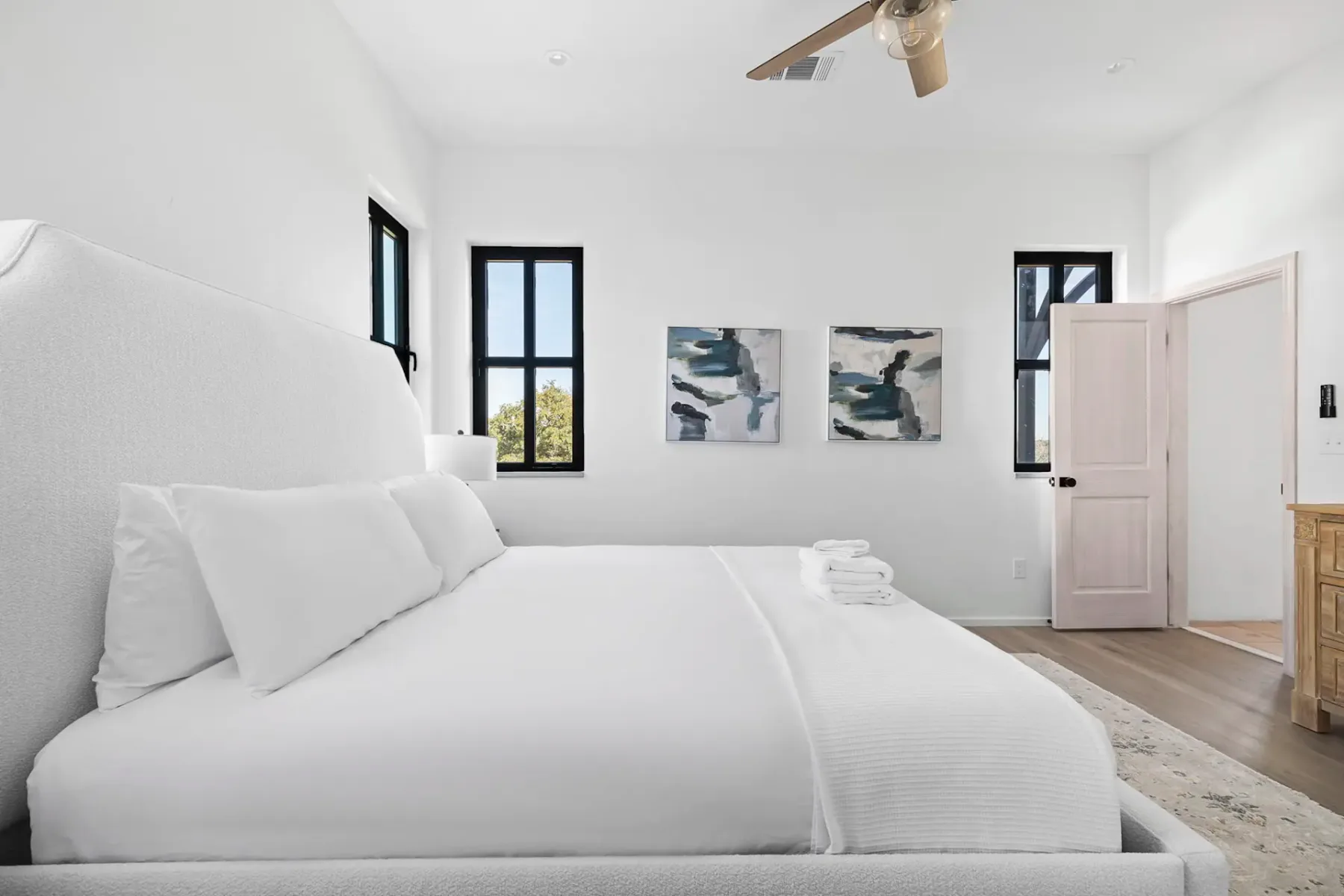Minimalist bedroom with a large white bed, abstract wall art, wooden dresser, and three windows. Ceiling fan and natural light enhance the airy feel.
