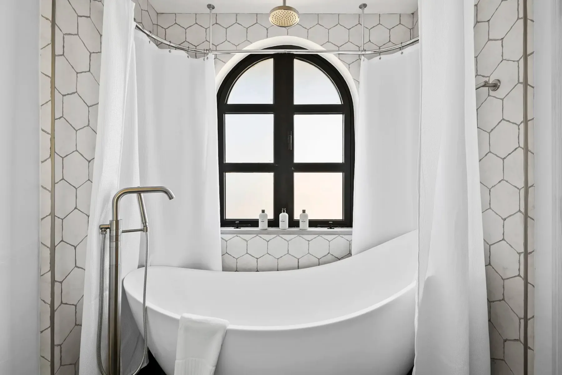 Modern bathroom with a white freestanding tub, hexagonal tile walls, a curved shower rod, and an arched window with bottles on the ledge.