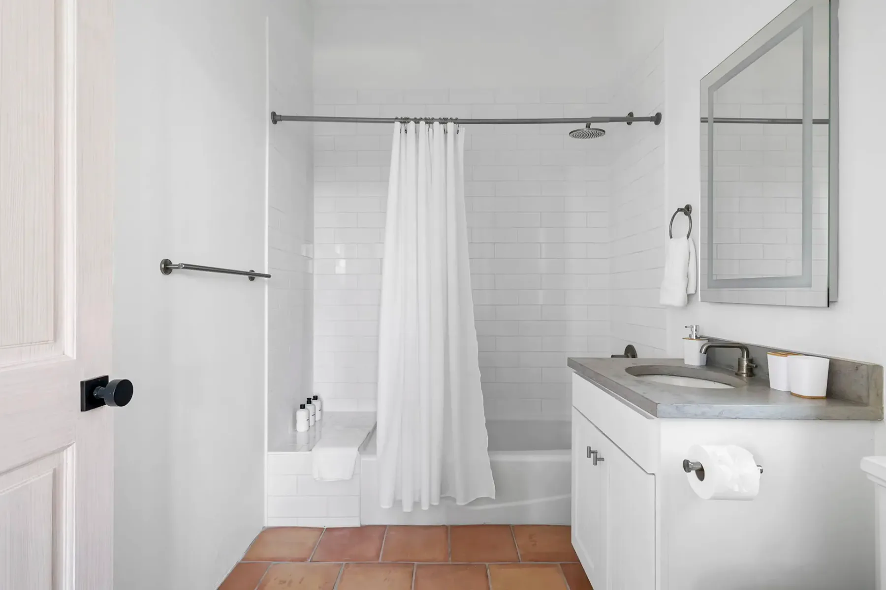 Minimalist bathroom with white tiled walls, a shower with a curtain, and a sink vanity. Terracotta floor tiles and a rectangular mirror complete the look.