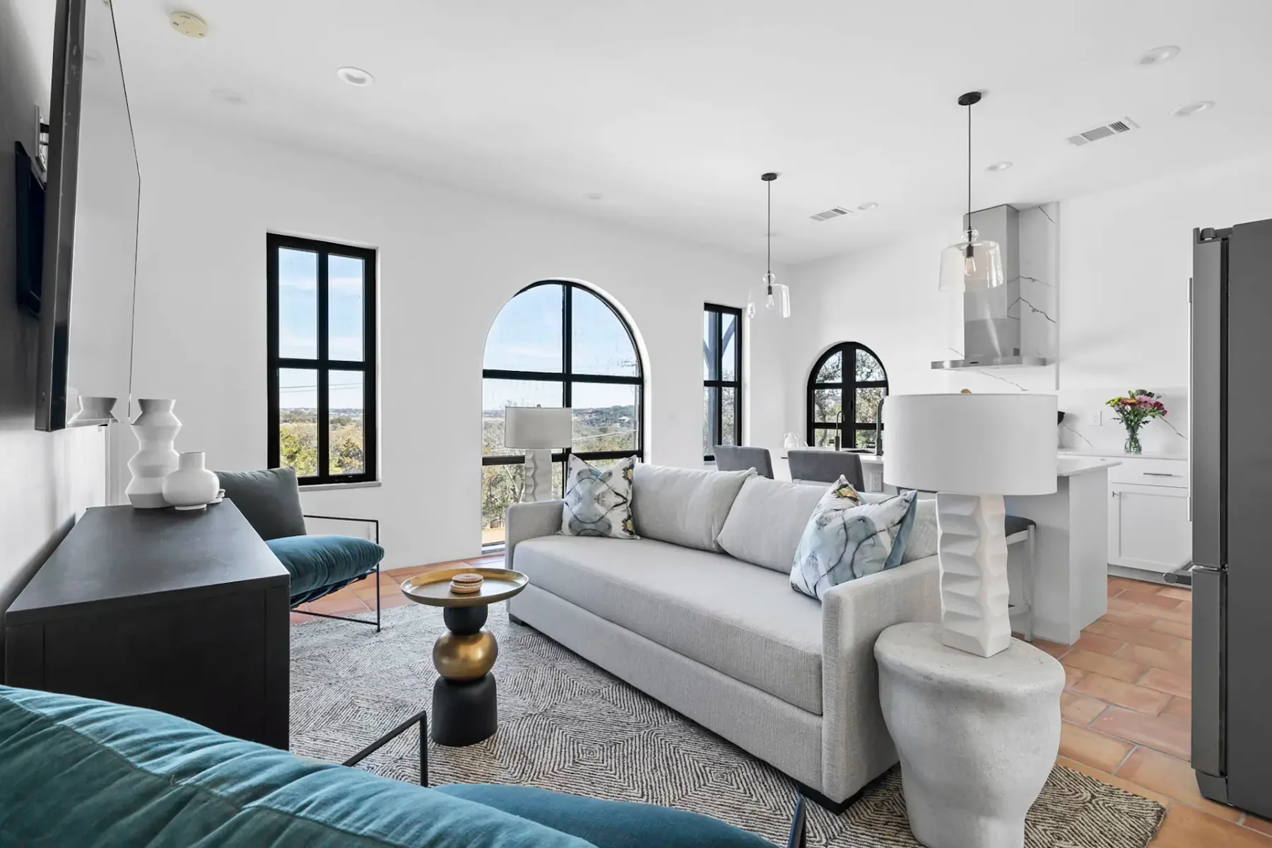 Modern living room with a gray sofa, blue chairs, abstract rug, arched windows, and a view of the kitchen with pendant lights in the background.